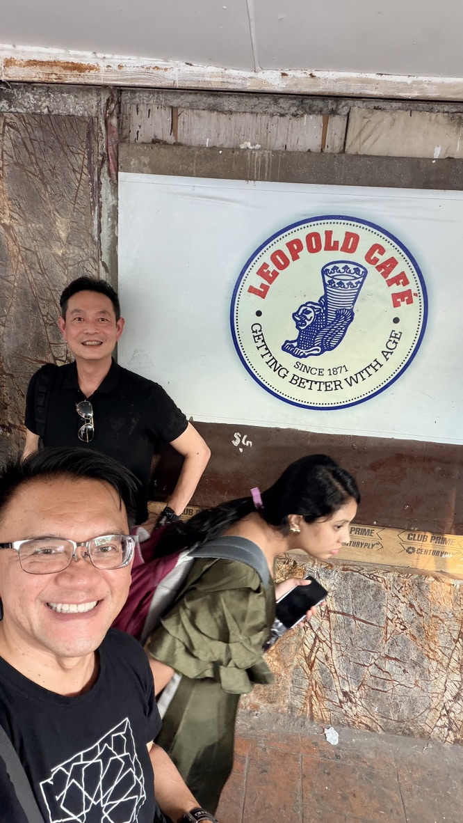 Two travellers beside the Leopold Café wall sign, Mumbai, India.