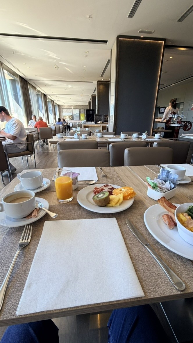 Coffee, orange juice, fruit, bacon, and breakfast plates on a table by the windows