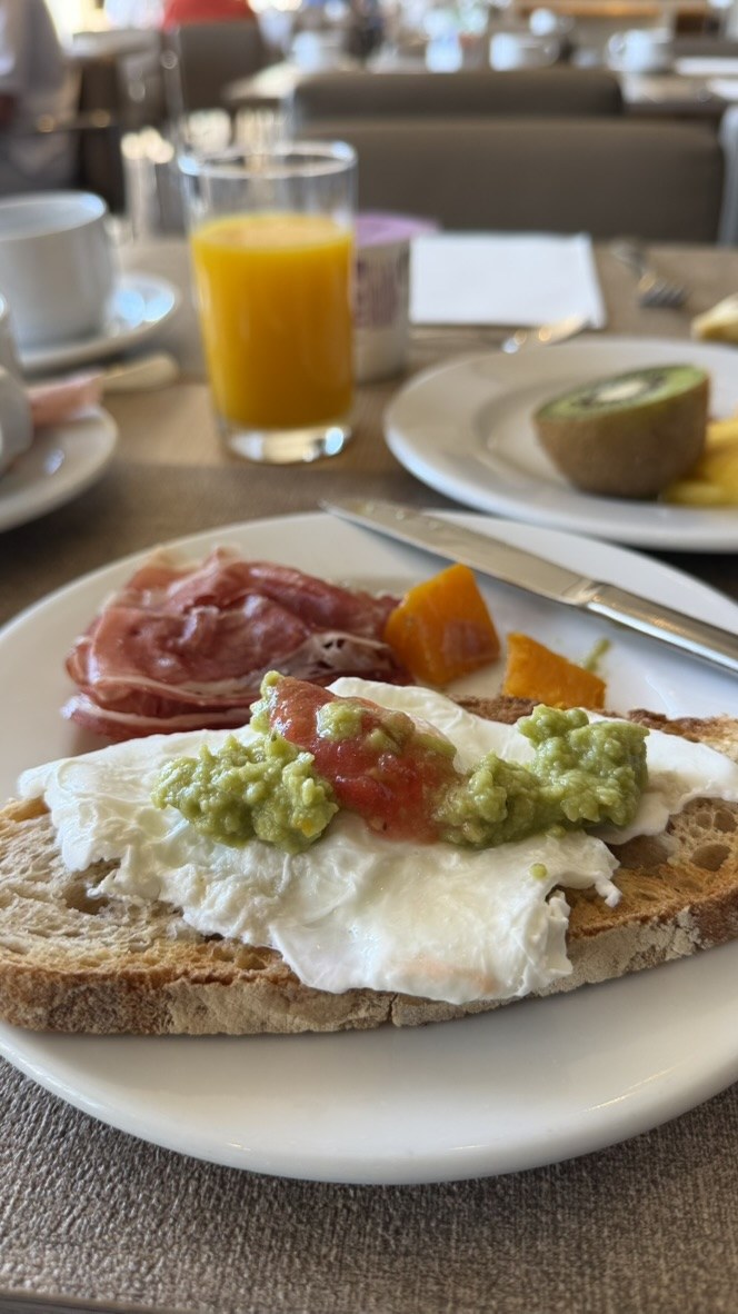 Rustic toast topped with egg, tomato, and avocado beside fruit and cured meat