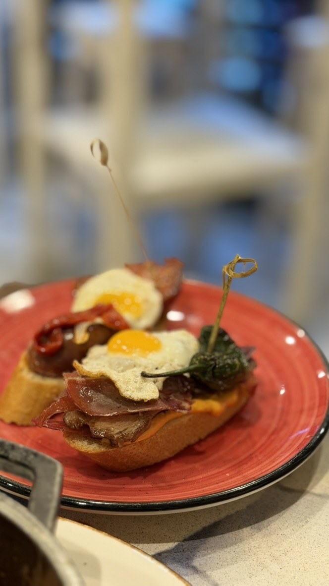 Two montaditos layered with cured meat, peppers, and fried quail eggs